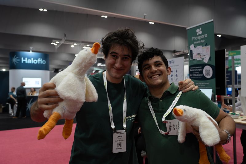 Leon Mojarrabi and Anis Mihrshahi posing with white plush geese on a trade-show floor, both wearing exhibitor badges